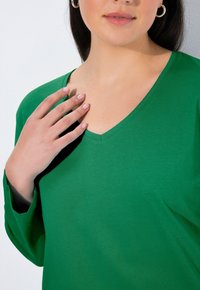 Zielona koszulka z długim rękawem i dekoltem w serek, wykonana z miękkiej bawełny. Charakteryzuje się swobodnym krojem i minimalnymi detalami szwów, podkreślającymi gładką fakturę.