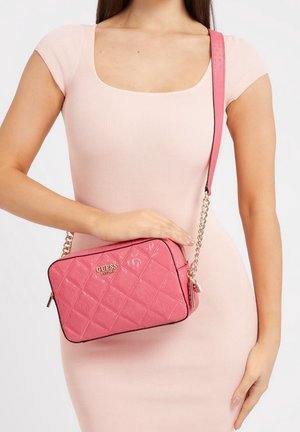 Femme en robe rose clair tenant un sac bandoulière matelassé rose Guess avec une chaîne dorée sur fond blanc uni.