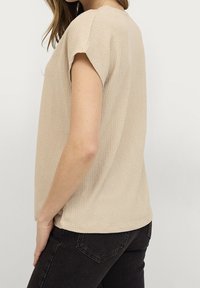 Beige textured top with short rolled sleeves, a relaxed fit, and a rounded hem. Paired with dark jeans, showcasing a subtle pattern.