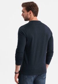 Camicia a maniche lunghe blu navy realizzata in tessuto morbido, con scollatura a girocollo e maniche aderenti. Indossata con jeans blu, che mostrano un design minimalista.