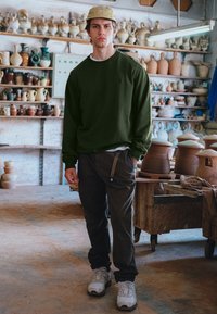 Sudadera verde oscuro de manga larga, combinada con pantalones oscuros que tienen una cinta de lado, y zapatillas blancas. Fondo neutro de cerámica.