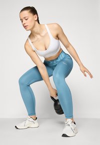 Sportski grudnjak je bijel, tajice su svijetloplave s "NIKE" logotipom. Model drži crnu girju, noseći bijele tenisice sa sivim akcentima.