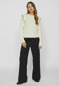 Kremowy sweter z falbaniastymi akcentami na ramionach, zestawiony z szerokimi czarnymi spodniami. Gładka tekstura, luźny krój i pełne rękawy.
