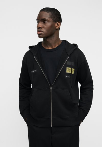 Μαύρη φούτερ ζακέτα με φερμουάρ, τσέπες και υφασμάτινη κουκούλα, φορεμένη πάνω από μαύρο μπλουζάκι. Διακριτικές λεπτομέρειες με επιδιορθώσεις στο στήθος.