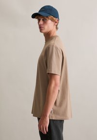 Beżowy bawełniany t-shirt z krótkimi rękawami, o luźnym kroju, z okrągłym dekoltem i bocznym rozcięciem. Nosi się go z czarnymi jeansami i niebieską czapką.