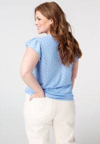 Haut bleu clair en dentelle à manches courtes et volantes. Le tissu présente un motif texturé, associé à un pantalon de couleur crème.