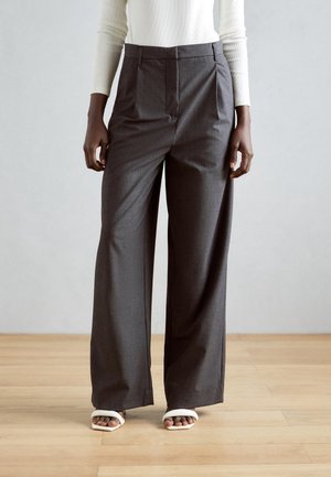 Persoon die donkere wijde broekspijpen, een witte top met lange mouwen en witte open-toe sandalen draagt, staand op een houten vloer.