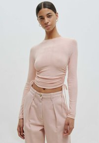 Langärmliges, geripptes Oberteil in zartem Rosa, mit gerafften Seiten und Bindebändern. Kombiniert mit hochgeschnittenen, taillierten rosa Hosen. Glatte Textur.
