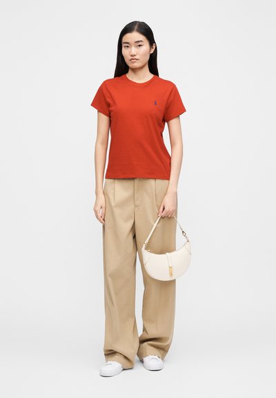 Piros pamut rövid ujjú póló kis kék logóval, bézs redőzött nadrággal és fehér íves táskával párosítva. Fehér sportcipő.