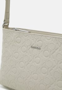 Kabelka Calvin Klein v světle béžové kůži s raženým vzorem loga CK. Disponuje zipovým uzávěrem a nastavitelným popruhem. Texturovaný povrch.