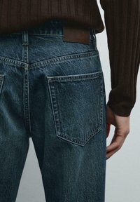Jeans in denim blu con vita alta, tasche posteriori e un'etichetta in pelle. Mano appoggiata sui fianchi, visibile una maglietta marrone texturizzata.