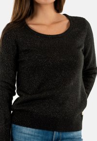 Un pull noir avec un décolleté en cœur, texture scintillante, manches longues et ourlet côtelé; associé à un jean en denim bleu. Présence de paillettes argentées brillantes tout au long.