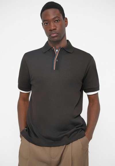 Μαύρο πόλο μπλουζάκι με πολύχρωμο ριγέ καπάκι, κοντά λευκά μανίκια, φτιαγμένο από λείο ύφασμα, φορεμένο με μπεζ παντελόνι.