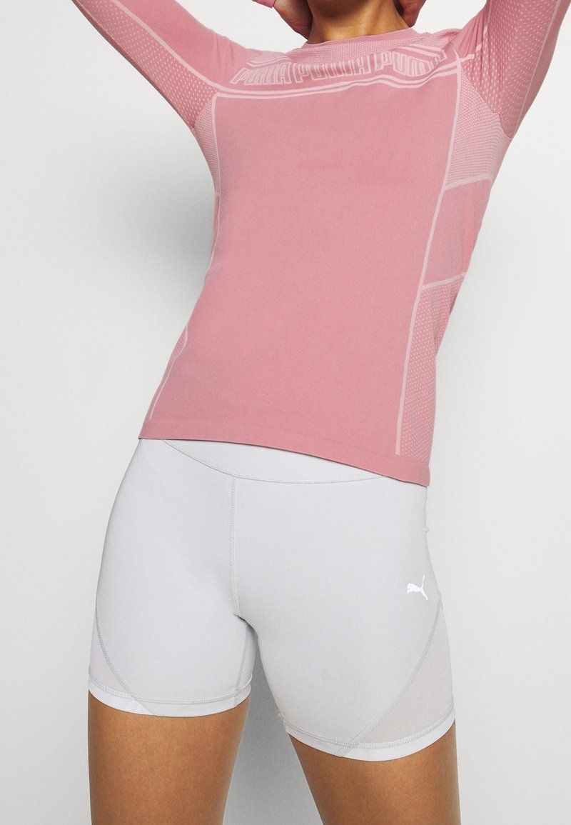 Světle růžové atletické tričko s dlouhým rukávem, texturovanými panely a logem, doplněné bílými přiléhavými šortkami s průsvitnými detaily a jemným prošitím.
