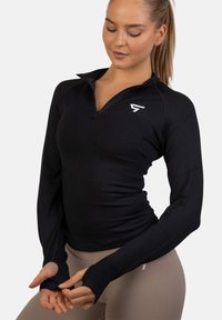 Crna dugorukava sportska majica s patent zatvaračem i uskim krojem, rupicama za palčeve i malim bijelim logom na prsima. Glatka tekstura tkanine.