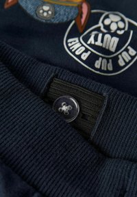 Tumši zilā ribota josta ar regulējamu melnu elastīgu materiālu un caurspīdīgu pogu, daļa apģērba ar „Pup Power Duty” suņu tematisko uzšuvumu.