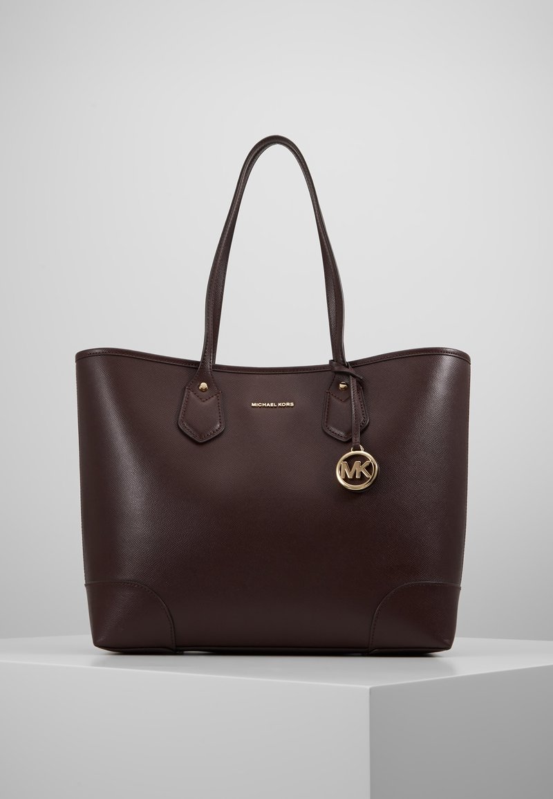 Sac cabas Michael Kors en cuir marron foncé avec double poignée et pendentif logo MK doré, présenté sur un socle blanc devant un fond gris.