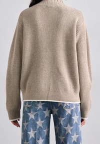 Person som bär en beige ribbad tröja och blå jeans med ett vitt stjärnmönster, visad bakifrån mot en enhetlig bakgrund.