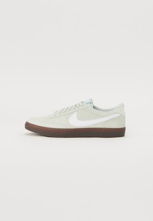 Zapatillas de ante gris con logo en forma de swoosh blanco, puntera redonda, cordones blancos y suela de goma marrón. Textura suave y diseño minimalista.