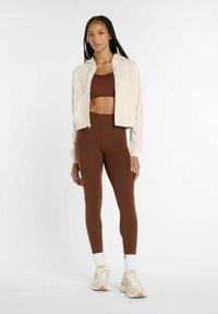 Krämfärgad zip-up hoodie, brunt croptop, bruna högmidjade leggings och beige sneakers med vita strumpor. Släta texturer, figurnära design.