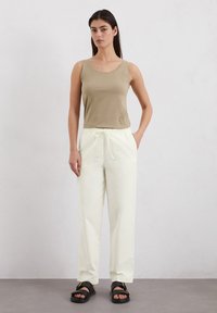 Beige Tanktop, entspannte weiße Hose mit Kordelzug, schwarze Sandalen mit metallischen Schnallenakzenten, vor einem neutralen Hintergrund.