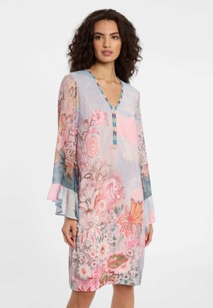 Frau trägt ein pastellfarbenes, knielanges Kleid mit Blumenmuster, langen Glockenärmeln und einer mit Perlen verzierten V-Ausschnitt, steht vor einem schlichten weißen Hintergrund.