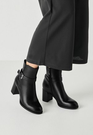 Person wearing black leather ankle boots with block heels and side buckles, paired with black wide-leg pants standing on a light floor.
