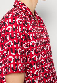 Homme portant une chemise rouge à manches courtes ornée d'un motif répétitif de texte noir et blanc "HUGO", debout devant un fond clair uni.