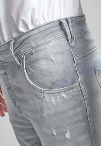 Jasnoszare dżinsy z efektem przetarcia, z postrzępionymi brzegami i widocznym przeszyciem. Zawierają przednie i tylne kieszenie oraz metalowe akcesoria.