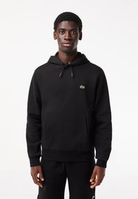 Jeune homme portant un sweat à capuche noir avec un petit logo crocodile vert sur la poitrine, debout devant un fond blanc uni.