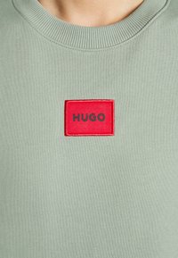 Felpa verde con scollatura a girocollo e un'etichetta rettangolare rossa con la scritta "HUGO" in testo nero. Tessuto morbido in tutto il capo.