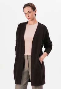 Brauner, flauschiger Cardigan mit offener Vorderseite und zwei seitlichen Taschen. Lange Ärmel und lockere Passform, kombiniert mit einem hellrosa Top und grauen Hosen.