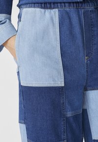 Džínové nohavice z patchworku s striedajúcimi sa tmavomodrými a svetlomodrými sekciami, s elastickým pásom a zvýrazneným šitím pre textúru.