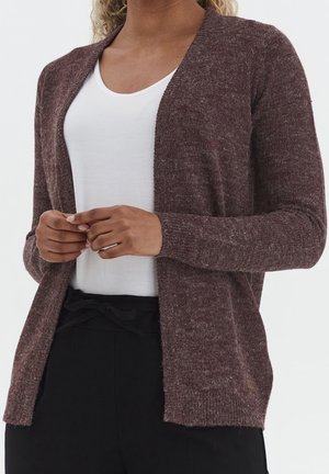Femme portant un cardigan marron texturé ouvert devant, sur un top blanc à encolure dégagée et un pantalon noir noué devant par un nœud.