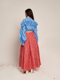 Blå bluse med flæser ved ærmerne og høj krave, parret med en lyserød broderet nederdel med blomstermønstre. Sandaler i beige med stropper er synlige.