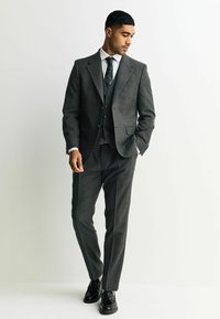 Traje de lana gris con un ajuste entallado, que presenta solapas de muesca y tres botones, usado sobre una camisa blanca y una corbata con un patrón, combinado con zapatos negros.