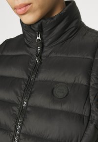Giacca nera imbottita con colletto alto, caratterizzata da una chiusura con zip e da una toppa rotonda con logo sul petto. Texture liscia, design leggero.