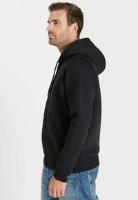 Zwarte hoodie met een voorkantzak, trekkoordkap en geribbelde boorden. Gecombineerd met lichtblauwe spijkerbroek, tegen een effen achtergrond.