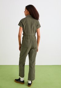 Tuta verde oliva con maniche corte, vita aderente e gambe dritte e morbide. Presenta tasche posteriori e tessuto strutturato.