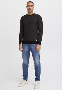 Svart strikket genser med rund hals, ribbestrikkede ermer og nederkant. Kombinert med lyseblå jeans og beige joggesko, stående mot en nøytral bakgrunn.