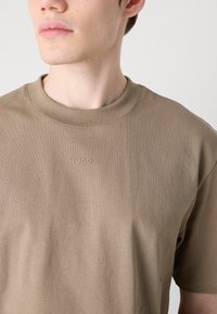 Khaki bavlnené tričko s krátkym rukávom a okrúhlym výstrihom, s jemne embosovaným logom. Tkanina má hladkú, mäkkú textúru.