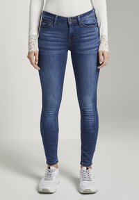 Mallas vaqueras ajustadas en denim azul oscuro, con un diseño clásico de cinco bolsillos y un ligero desgaste. Llevadas con zapatillas blancas y una camiseta de manga larga.