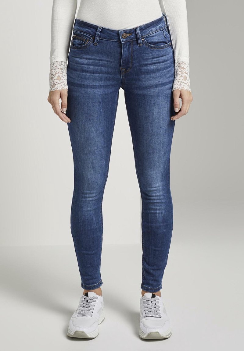 Mallas vaqueras ajustadas en denim azul oscuro, con un diseño clásico de cinco bolsillos y un ligero desgaste. Llevadas con zapatillas blancas y una camiseta de manga larga.