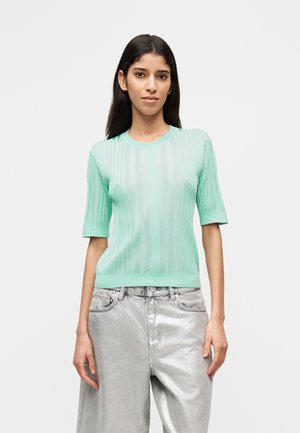 Kobieta o długich ciemnych włosach ubrana w miętowo-zieloną prążkowaną bluzkę z krótkim rękawem oraz metalicznie srebrne, luźne jeansy z wysokim stanem na białym tle.