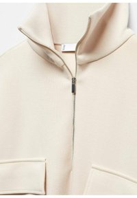 Sudadera beige con cremallera y cuello alto, confeccionada en tela suave y con dos bolsillos delanteros de parche. Se muestran detalles de cremallera metálica plateada.