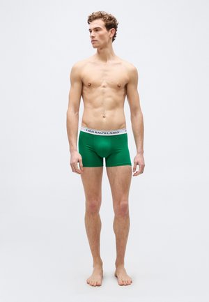 Jauns vīrietis stāv basām kājām, valkājot zaļas bokserbikses ar baltu jostas daļu, uz kuras rakstīts "Polo Ralph Lauren", skatoties pa kreisi.