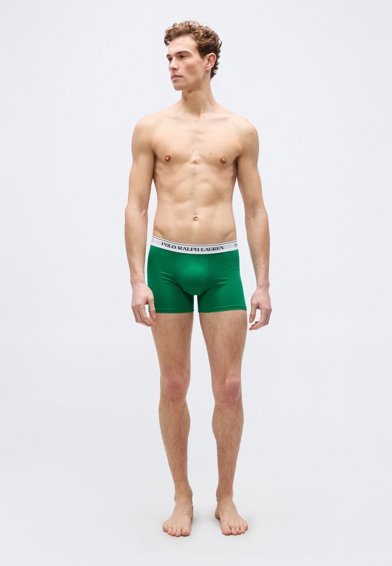 Νεαρός άνδρας που στέκεται ξυπόλητος, φορώντας πράσινα μποξεράκια με λευκή μέση που φέρει την ετικέτα "Polo Ralph Lauren", κοιτάζοντας προς τα αριστερά του.