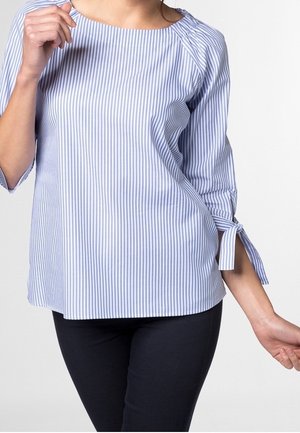 Frau trägt eine hellblaue und weiße Bluse mit vertikalen Streifen und dreiviertel Ärmeln, die mit Schleifen gebunden sind, kombiniert mit dunkler Hose.