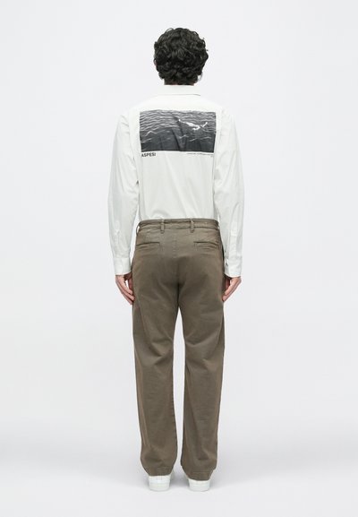 Λευκό πουκάμισο με μακριά μανίκια και σχέδι ο στο πίσω μέρος που απεικονίζει κυματισμούς νερού και κείμενο, σε συνδυασμό με καφέ παντελόνι και λευκά αθλητικά παπούτσια. Λείο ύφασμα.