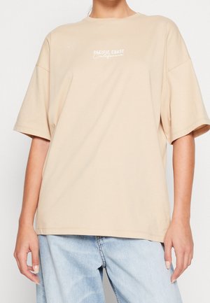 Beige oversized t-shirt made of soft cotton, featuring a white printed logo "PACIFIC COAST California" centered on the chest.